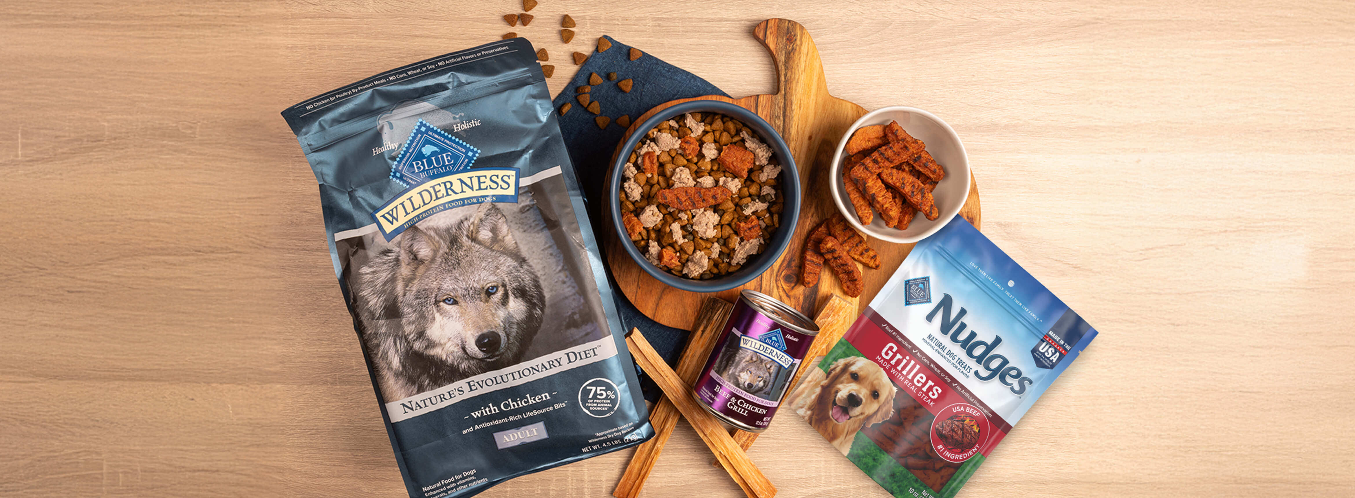 A dog-friendly meal sits in a bowl on a cutting board, surrounded by packages of BLUE Wilderness dry dog food, BLUE Wilderness wet dog food, and BLUE Nudges Grillers.