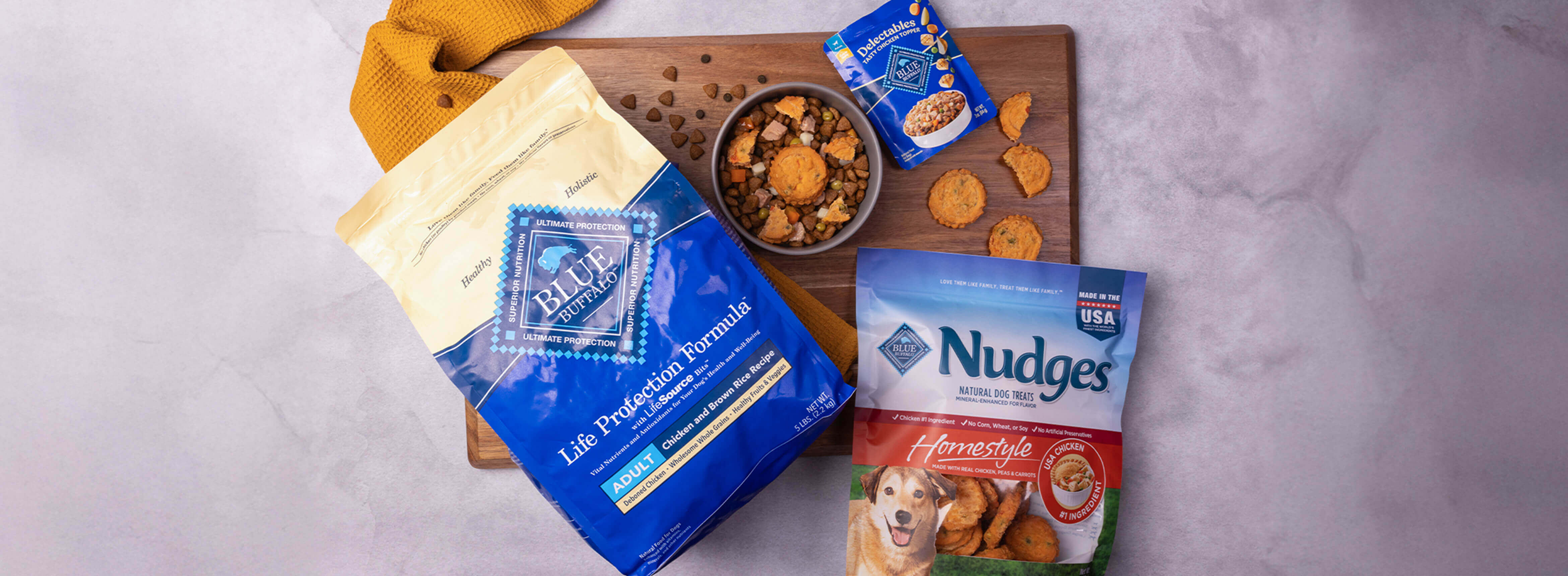 A dog-friendly meal sits in a bowl on a cutting board, surrounded by packages of BLUE Life Protection Formula, BLUE Delectables, and BLUE Nudges.