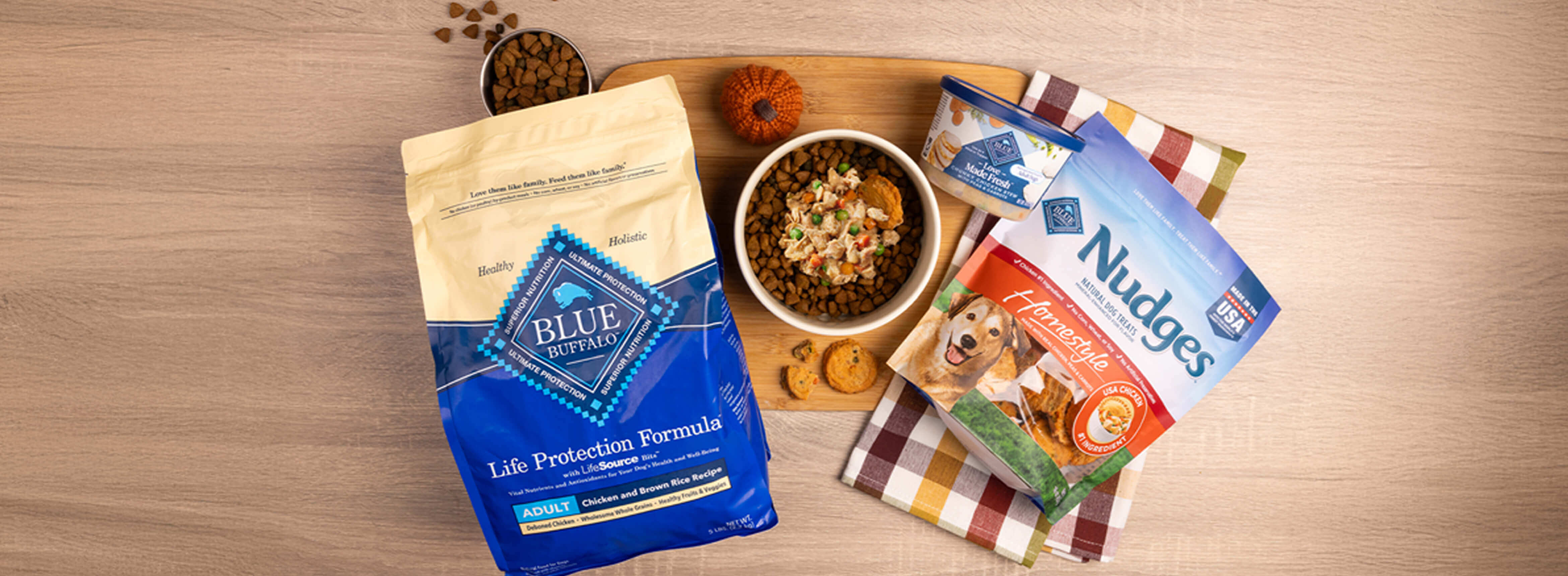 A dog-friendly meal sits in a bowl on a cutting board, surrounded by packages of BLUE Life Protection Formula, BLUE Nudges, and a tub of BLUE Love Made Fresh.